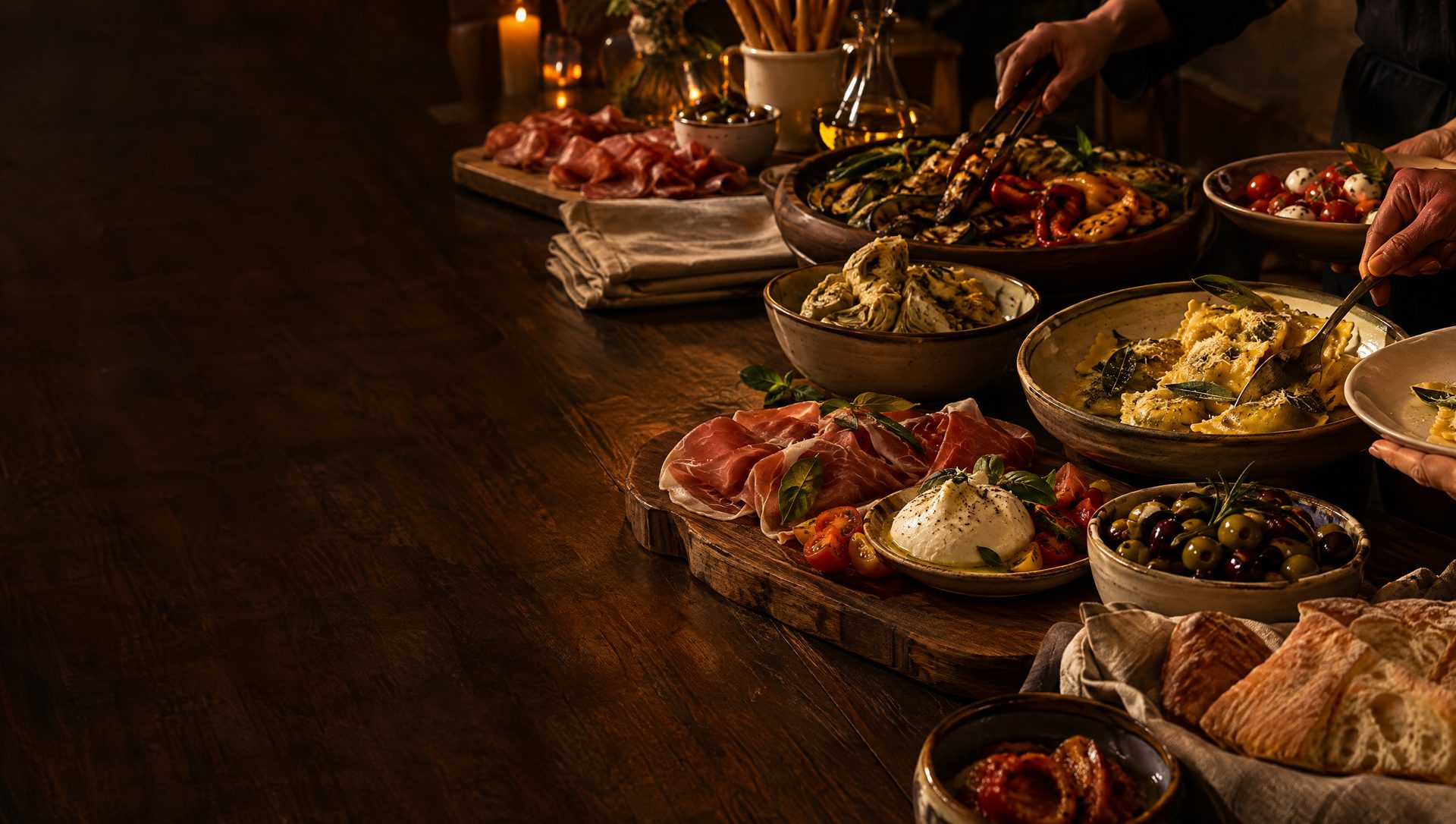Gedekte houten tafel met Italiaanse gerechten: antipasti, verse ravioli, prosciutto, burrata, olijven en ciabatta bij kaarslicht